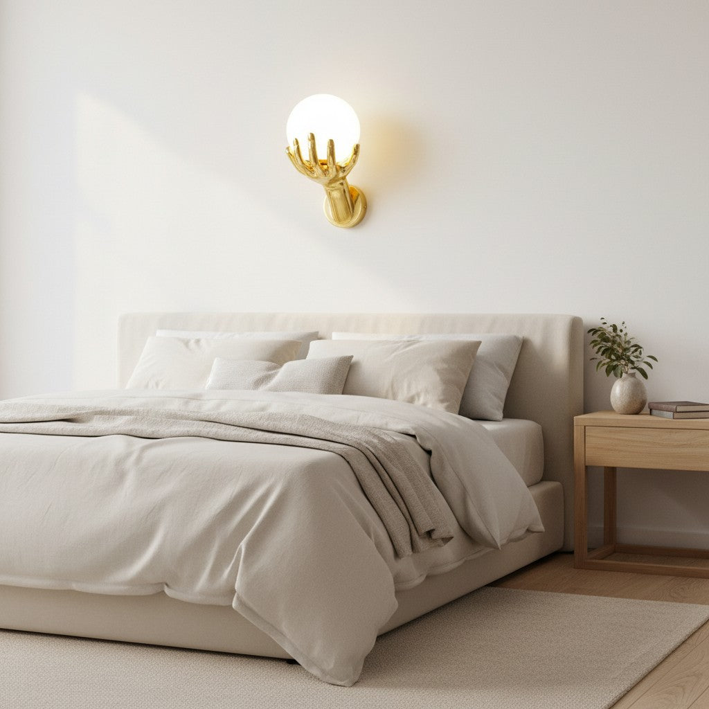 GoldenWatt gold hand-shaped wall lamp with a glowing frosted glass sphere bulb mounted above a modern neutral bedroom bed