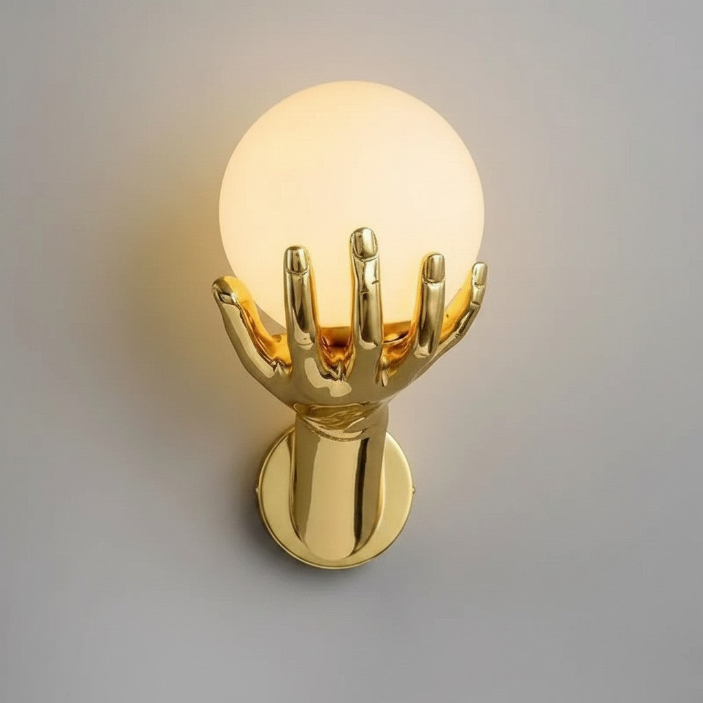 GoldenWatt gold wall sconce featuring a hand sculpture holding a glowing frosted glass orb for bedside lighting.